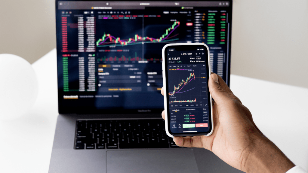 A person using their smartphone to view stock market data on their laptop.