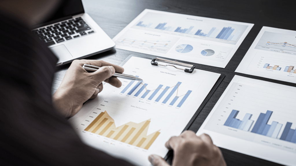 A hand is holding a pen and a calculator on a desk with papers and a laptop.